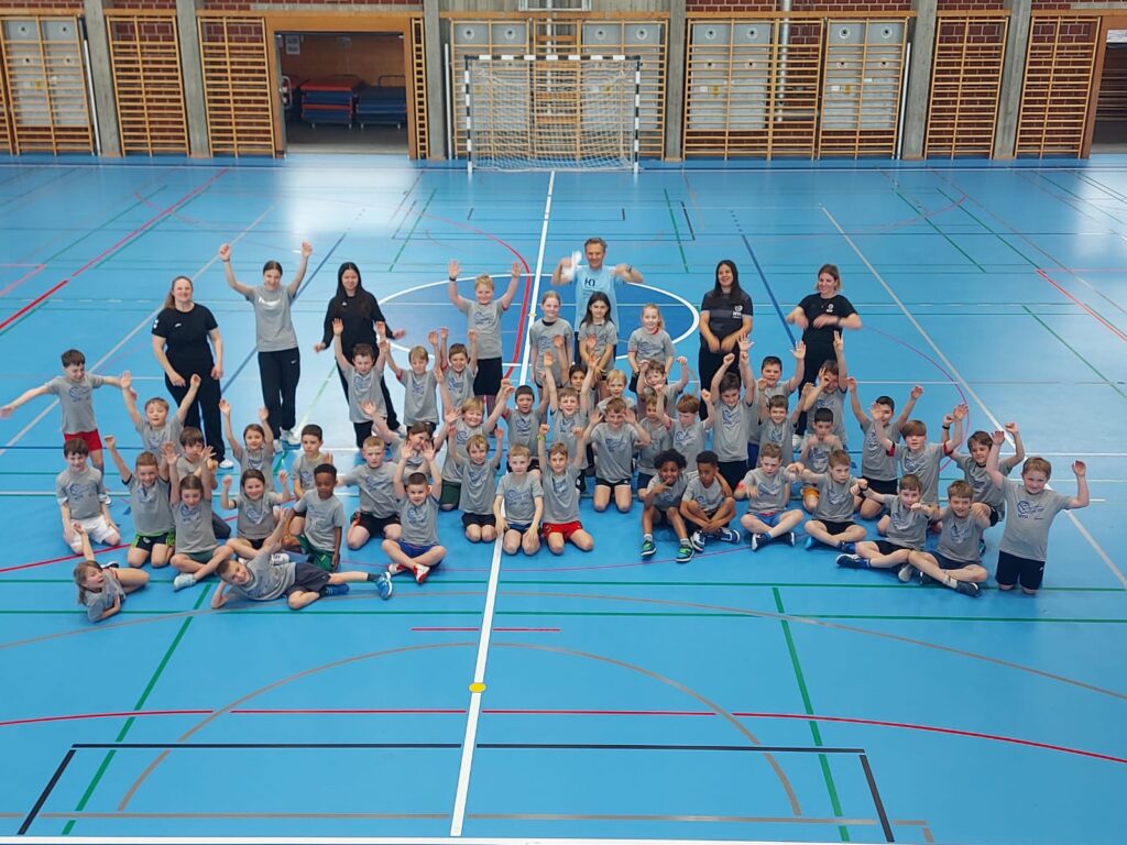 Large group of children in gray shirts posing for a team photo in a blue gymnasium, with coaches at the sides and hands raised in celebration.
