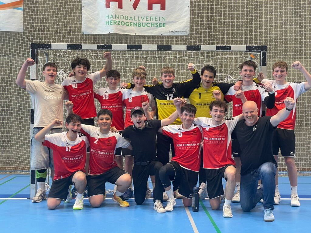 Group of young handball players posing triumphantly in front of a goal net inside a gym, wearing red and white uniforms.