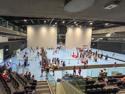 Indoor sports hall with many children playing on a blue court while spectators sit in the stands above and around.ériques