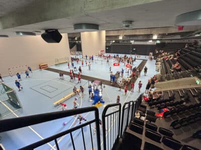 Indoor arena filled with kids participating in a sports event on a blue court, with spectators in the stands along the sides.
