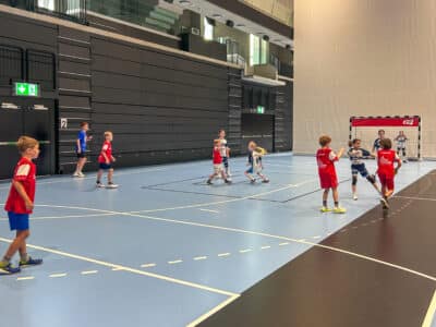 Youth handball game indoors with kids in red and blue uniforms on a blue court, a goal to the right and dark wall backdrop.