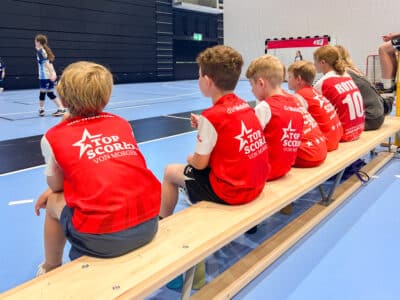 Young players in red jerseys sit on a bench along the sideline, watching a handball game in a gymnasium.