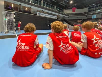 Young players in red jerseys sit on a blue indoor court, watching a handball match; a goalkeeper practices at the net in the background of a stadium.