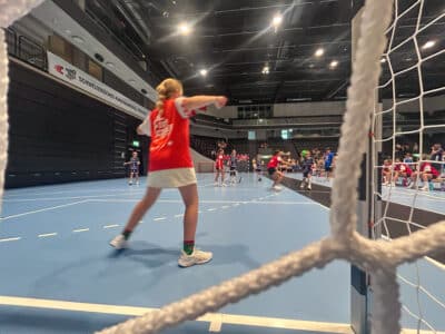 Indoor handball game in a arena; players in red jerseys near the goal as spectators watch from the stands, banner in the background reading 'Schweizerisches Kinderhandball-Festival'