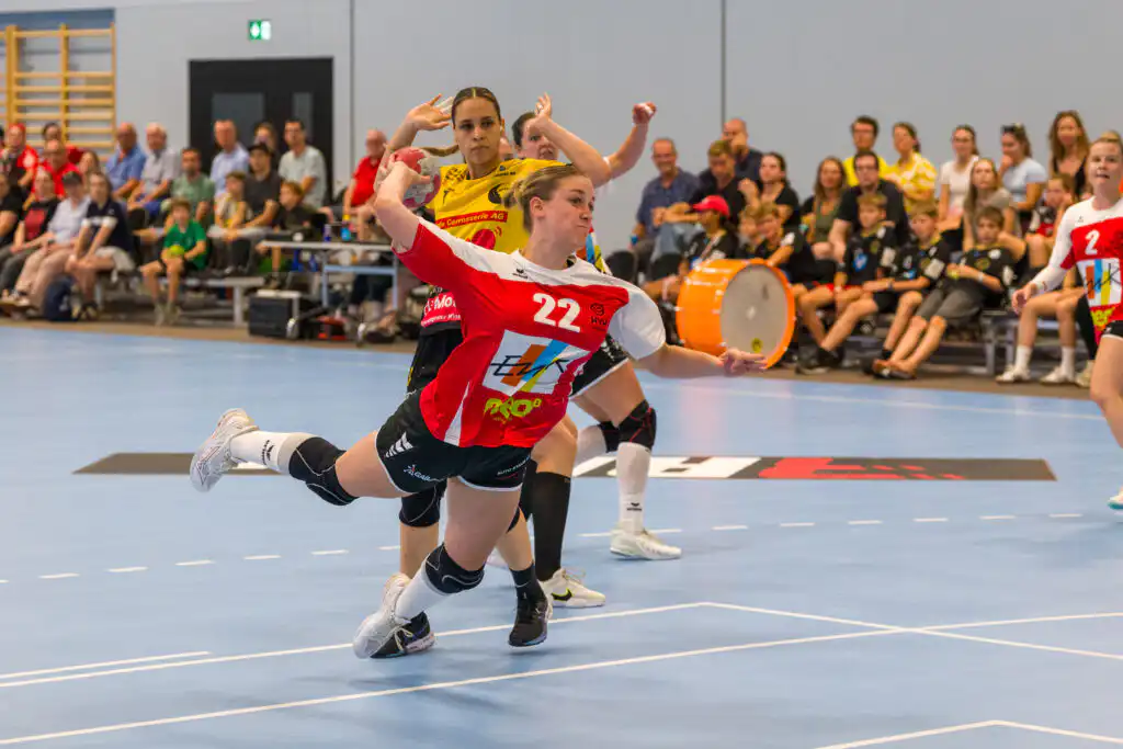 Handball player in red and white jersey #22 lunges forward with the ball on a blue court, spectators watching in the background.