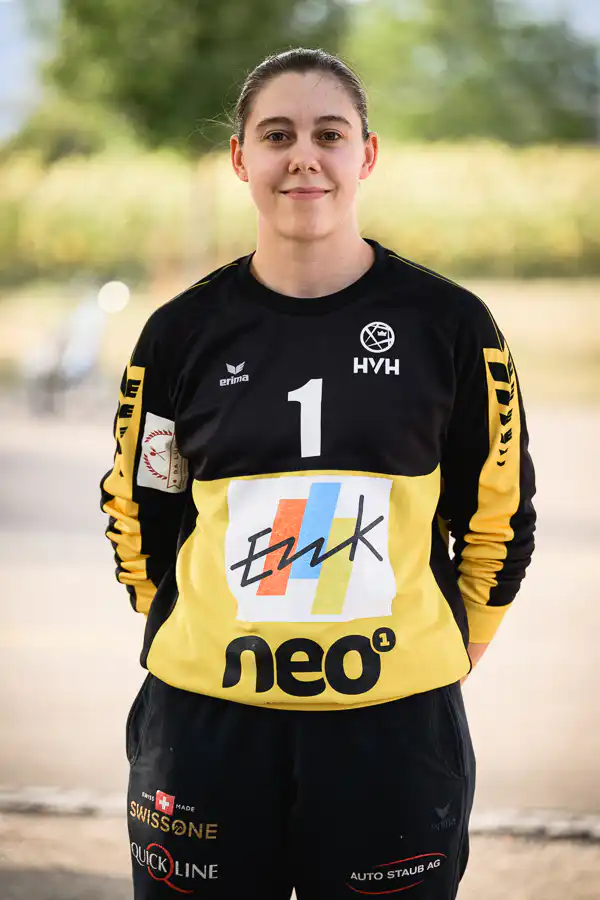 Female athlete standing outdoors in a black and yellow jersey with the number 1 and sponsor logos, smiling at the camera.