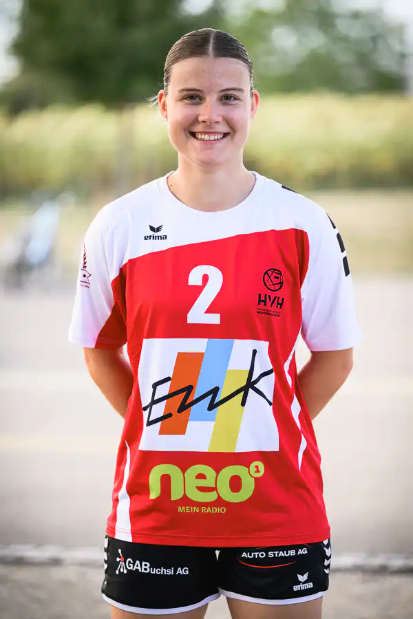 Smiling female athlete in a red-and-white jersey with the number 2 and multiple sponsor logos, standing outdoors.