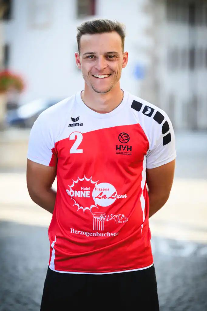 Smiling man in a red-and-white sports jersey with the number 2, standing outdoors with a blurred city street background.