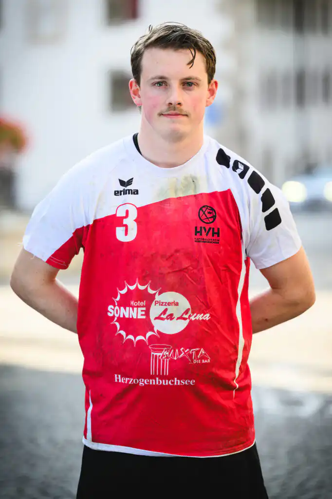 Young man in a red-and-white handball jersey with the number 3, standing outdoors near a waterway.