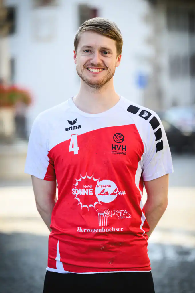 Smiling man in a white-red sports jersey with the number 4, standing outdoors with a blurred urban background.