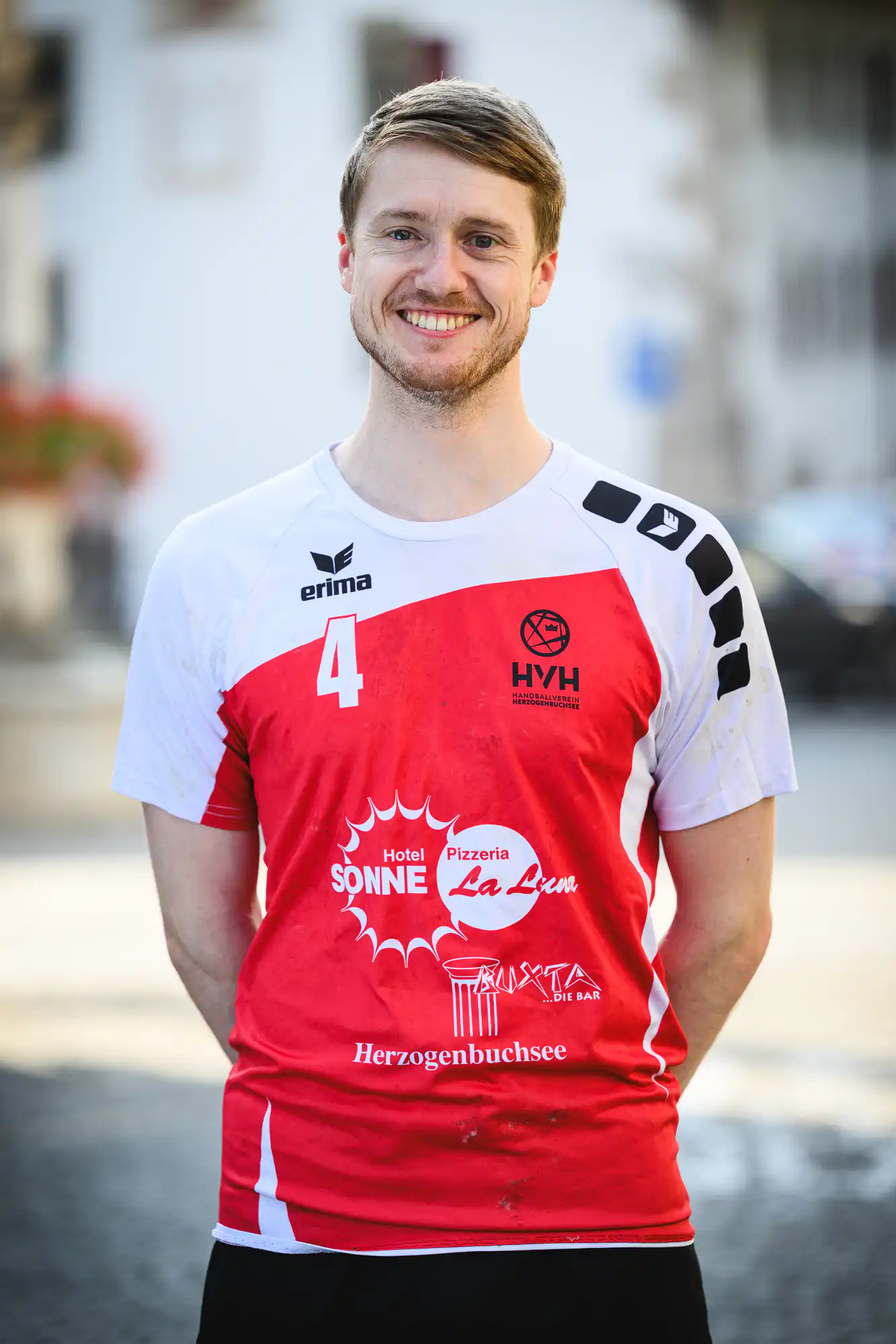 Smiling man in a white-red sports jersey with the number 4, standing outdoors with a blurred urban background.