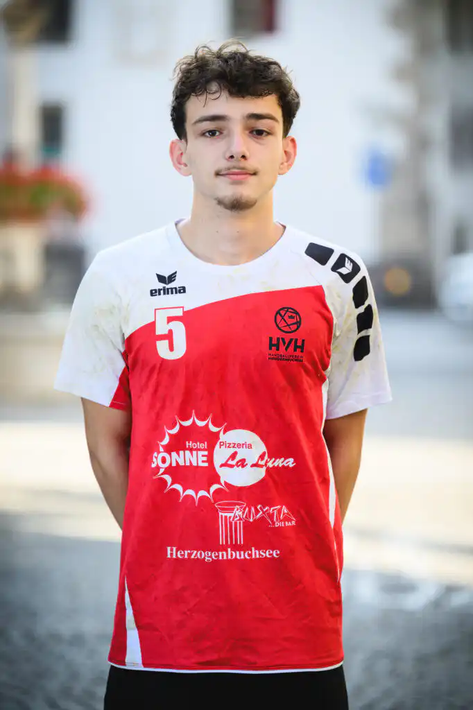Portrait of a young man in a red-and-white sports jersey with the number 5, standing outdoors near a waterway and looking at the camera.