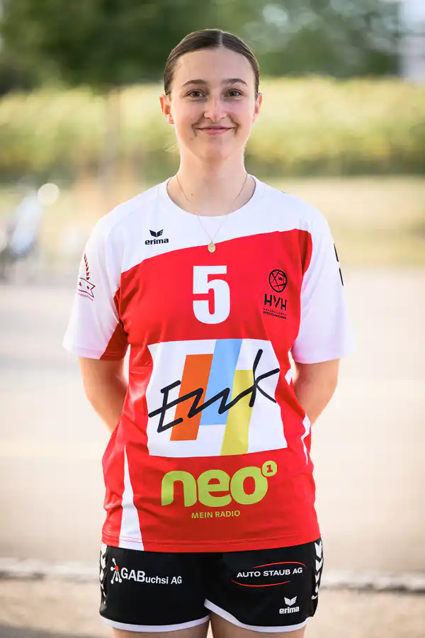 Young female athlete in a red-and-white jersey with number 5, standing outdoors and smiling.