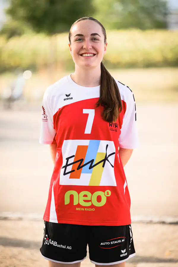 Smiling female volleyball player standing outdoors in a red-and-white jersey with number 7 and sponsor logos.