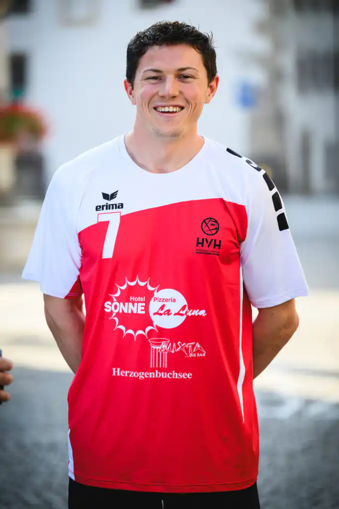 Smiling man in a white-and-red sports jersey standing outdoors, sponsor logos visible on the chest and torso.