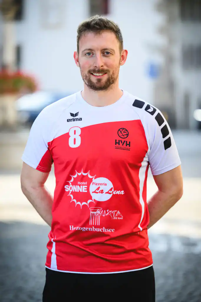 Man in a red-and-white sports jersey with the number 8, smiling, standing outdoors on a sunny street.