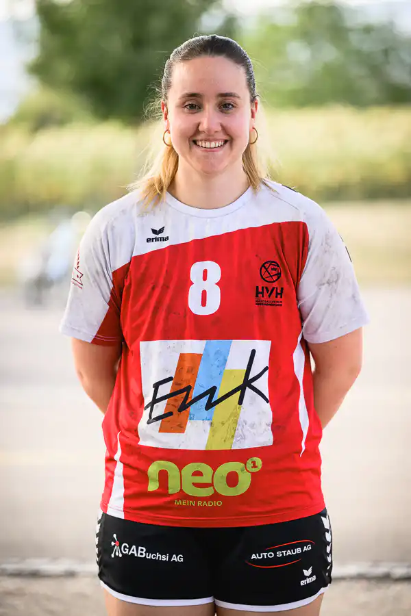 Female athlete standing outdoors in a red-and-white jersey with the number 8 and sponsor logos, smiling at the camera.