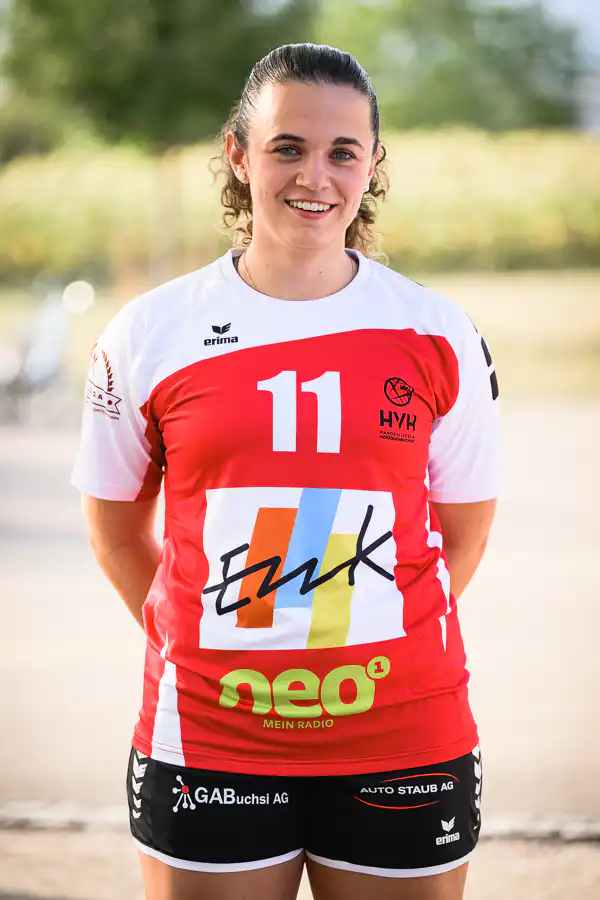 Female athlete in a red-and-white jersey with number 11, smiling outdoors, arms behind her back.