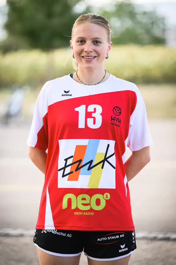 Young woman athlete standing outdoors in a red-and-white sports jersey with the number 13 and sponsor logos on the chest.