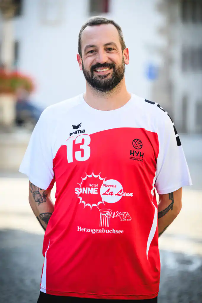 Portrait of a man in a red-and-white sports jersey with number 13, smiling outdoors.
