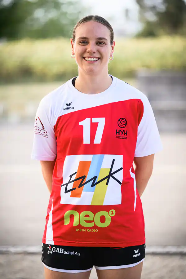 Female athlete in a red-and-white jersey with the number 17, smiling outdoors. The shirt features sponsor logos and a colorful 'tk' graphic.