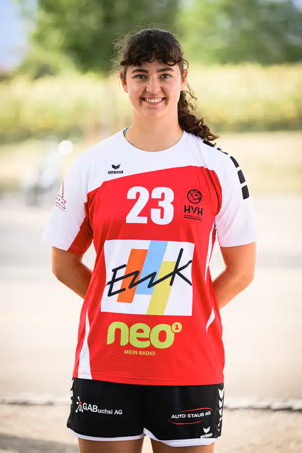 Female athlete in a red-and-white jersey with number 23, standing outdoors and smiling; sponsor logos visible on the shirt’s front and sleeves.