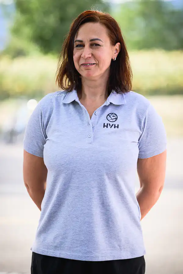 Portrait of a smiling woman with shoulder-length brown hair wearing a light gray polo with an HYH logo, outdoors.