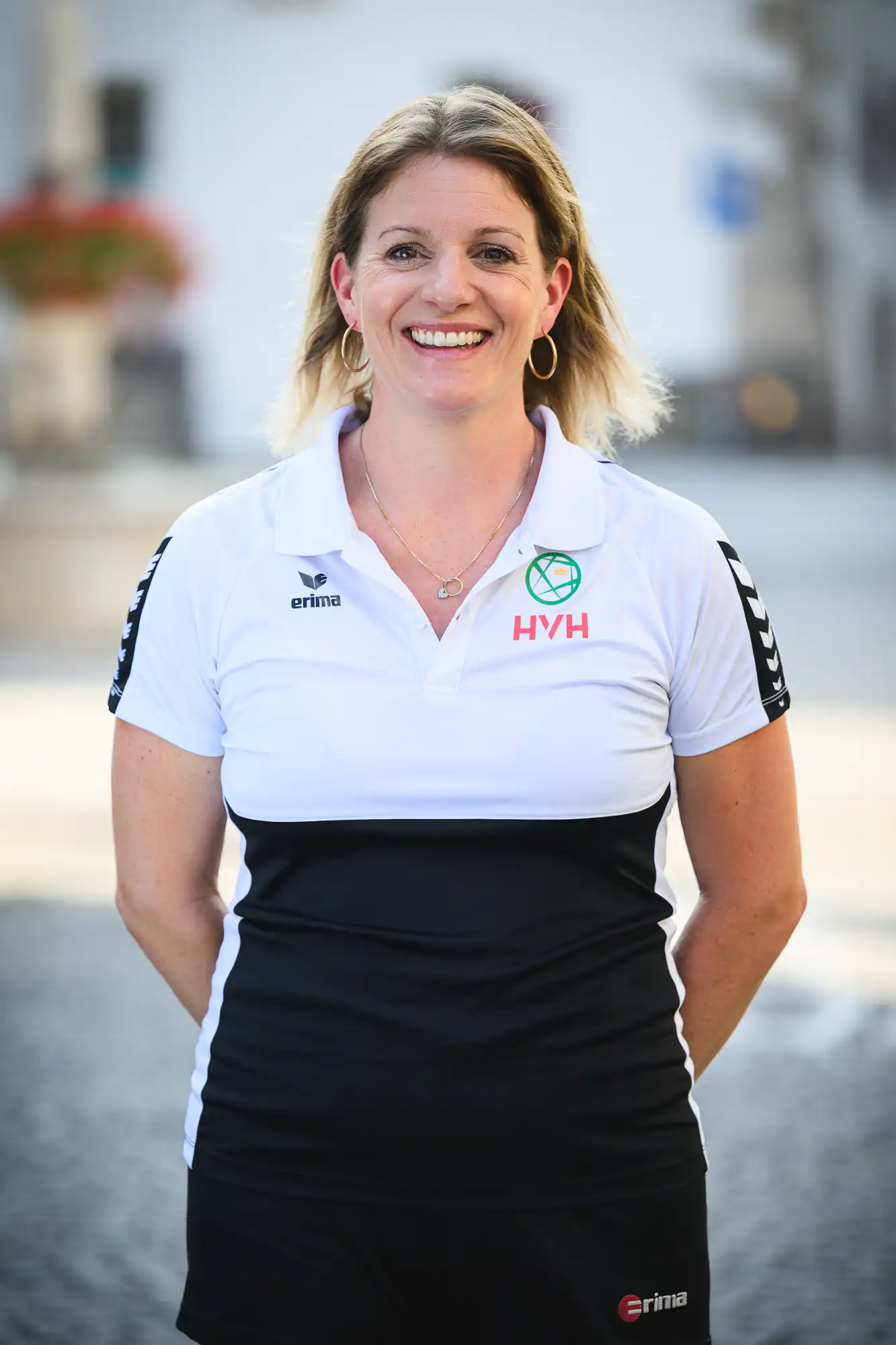 Smiling woman in a white polo shirt with HVH and Erima logos, standing outdoors.