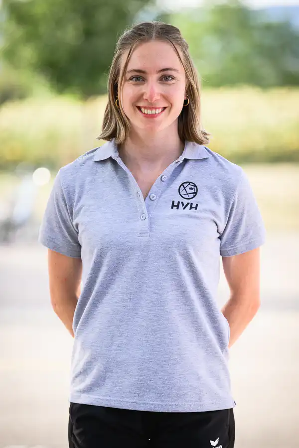 Smiling young woman standing outdoors in a light gray polo with a globe logo and 'HVH' on the chest.