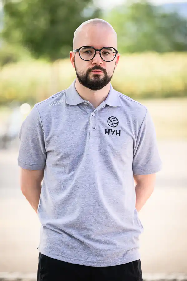 Man with a beard and black-ramed glasses standing outdoors in a light gray polo with an HVH logo on the chest.