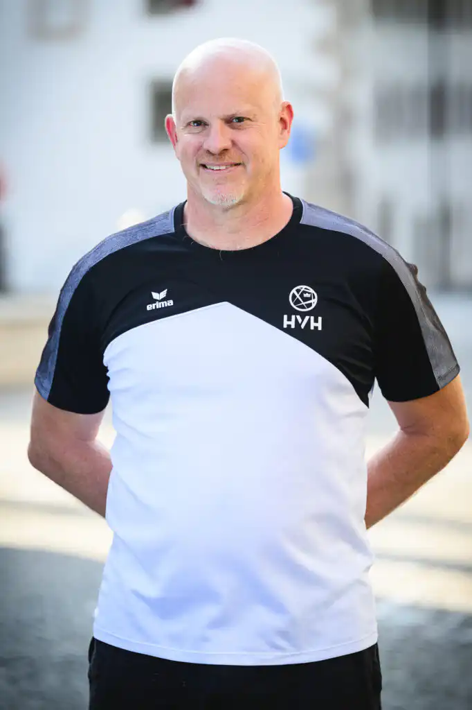 Smiling bald man in a black and white HVH sports shirt, standing outdoors.