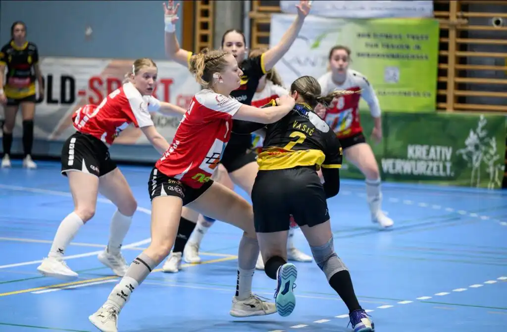 Women’s handball game in progress as red/white and black/yellow players collide near center court on a blue indoor court with banners in the background.