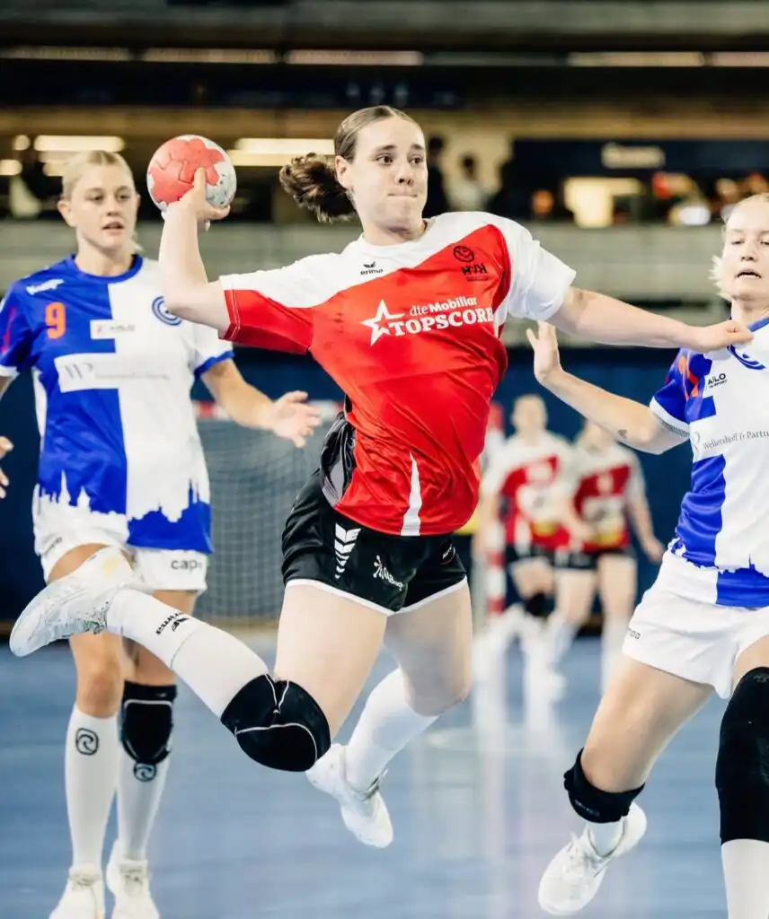 Female handball player in red jersey leaps with ball raised, aiming for goal during a game in an indoor arena, surrounded by opposing players in blue and white.