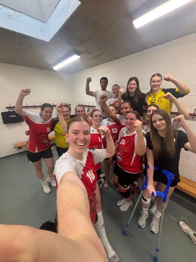 A group of female athletes in red and white uniforms celebrate with raised fists in a locker-room selfie, smiling at the camera.