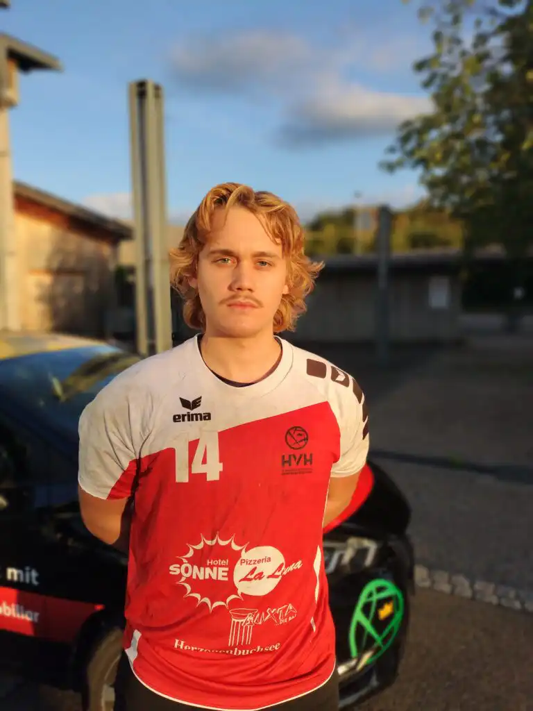 Young man with blond hair in a red-and-white sports jersey #14, standing outdoors with a blurred urban background.