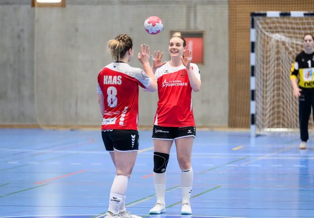 Two female handball players in red uniforms high-five on an indoor court after a play, ball in midair above them, with a goalie visible near the goal in the background.