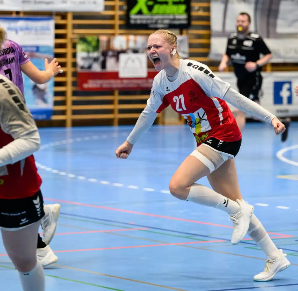 Female handball player in white and red jersey (number 27) sprinting and yelling on an indoor court during a match.