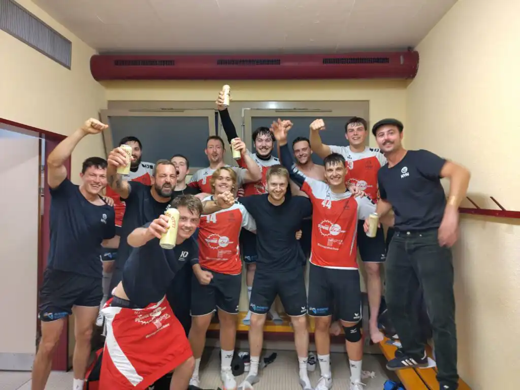 A group of male athletes in red and white jerseys posing in a locker room, cheering and holding up cans in celebration.