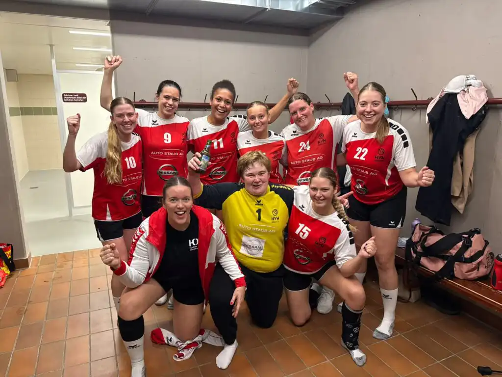 Girls' volleyball team in red and white uniforms cheer in a locker room, fists raised in celebration.